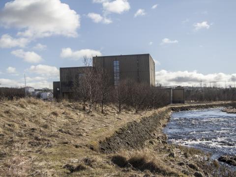 Toppstöðin in Elliðaárdalur, with yellow markings and the Elliðaá River in the foreground and a blue sky with scattered clouds in the background.