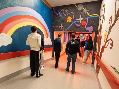 Young people seen from behind in a school hallway, surrounded by colorful walls, including a rainbow painted on one wall.