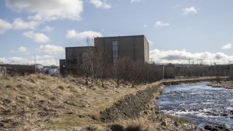 Toppstöðin in Elliðaárdalur, with yellow markings and the Elliðaá River in the foreground and a blue sky with scattered clouds in the background.